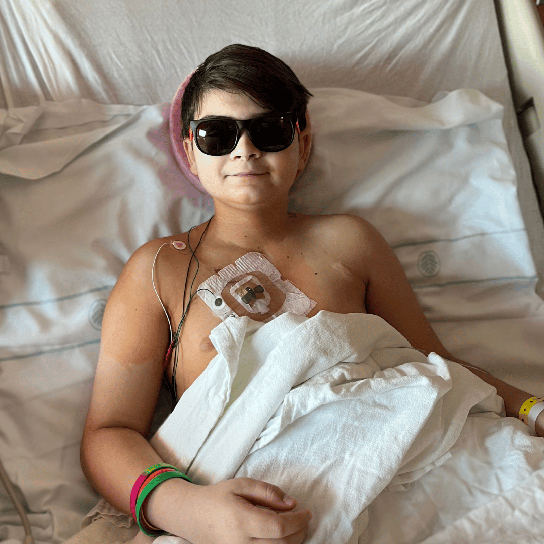 River, a boy with acute lymphoblastic leukemia wearing black sunglasses in his hospital bed with wires around his port