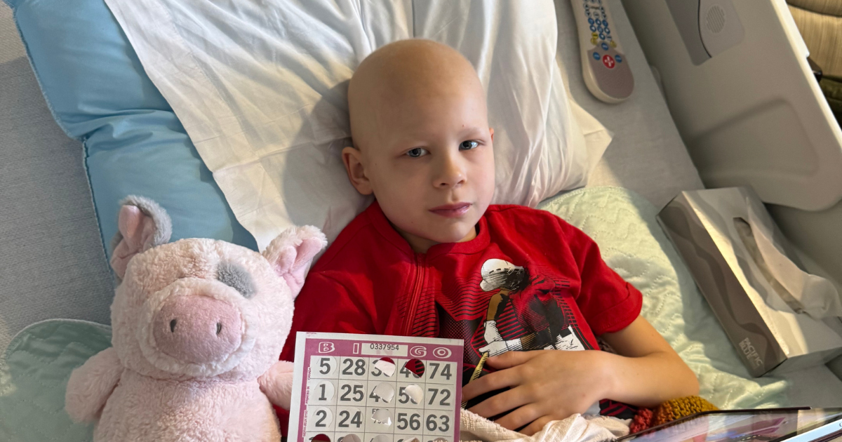 A little boy in a hospital bed with a stuffed pig sitting next to him while he holds up a bingo card.