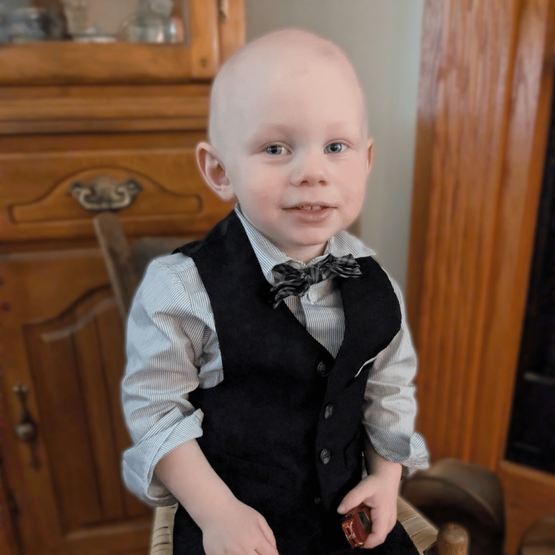 A photo of a little boy bald from cancer treatment wearing a tuxedo with a bowtie smiling at the camera.