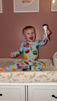 All-Star Lydia sitting on a changing table in a Sesame Street onesie holding a tube of lotion in the air. 