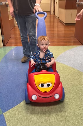 All-Star Lydia riding in a red toy car, being pushed by an adult in the hospital. 