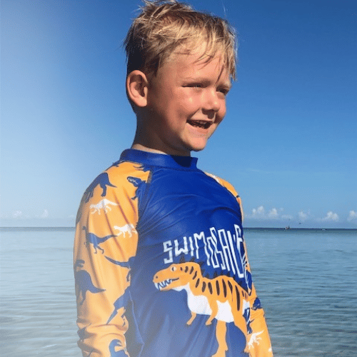 All-Star Eli squinting into the sun while wearing a swimming shirt with a dinosaur on it at a body of water.