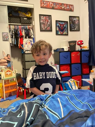 A child wearing a Baby Dude shirt standing by a bed
