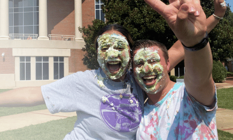 Two college students with pie on their faces smiling and giving a peace sign after a Cancer is Messy fundraiser to support kids with cancer.