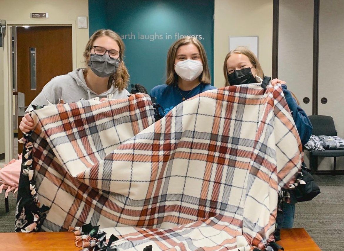 three college students holding a blanket for kids with cancer wearing masks.