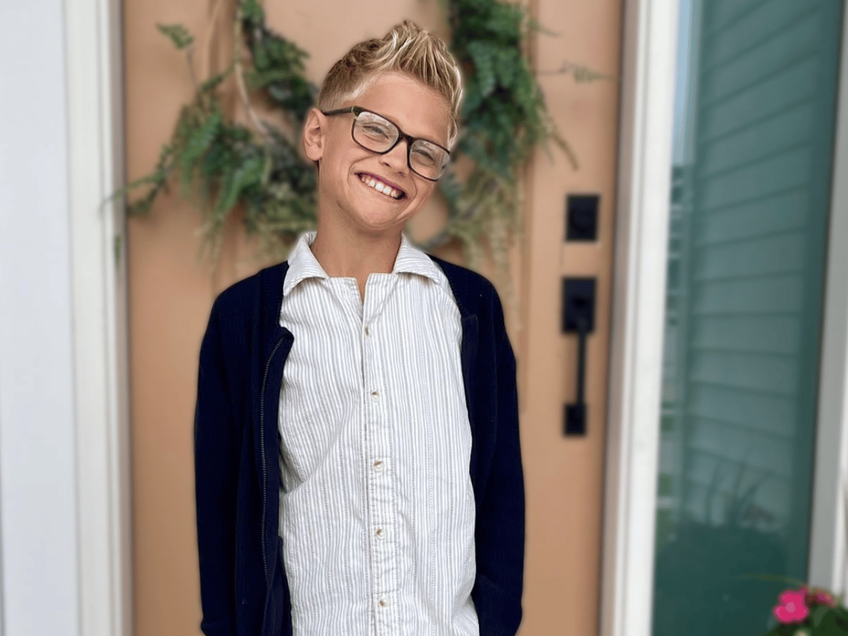 A little boy named Sage standing in front of a tan door wearing glasses and a button up shirt