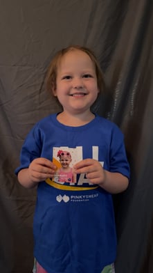 All-Star Scarlett holding a baseball card with her name on it after being featured by Pinky Swear Foundation when her family received financial support during her cancer diagnosis.