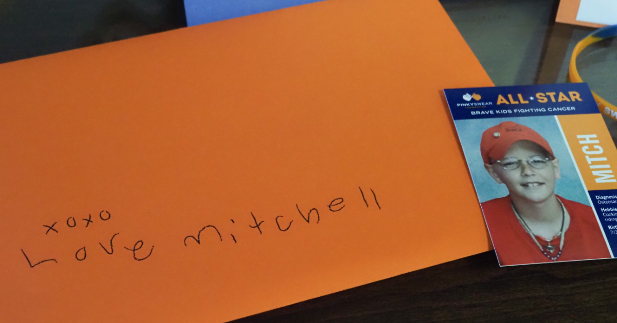 An orange envelope with the words "XOXO Love Mitchell" on it with a baseball card next to it with Pinky Swear Foundation's founder, Mitch on the front. Over 20 years ago, Mitch made his dad pinky promise to help other kids with cancer after he was gone. Pinky Swear supporters keep this pinky promise