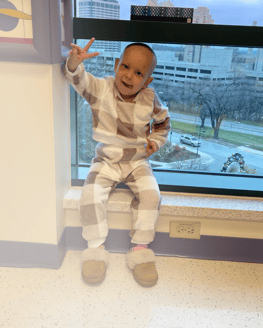 All-Star Kaylee sitting in the window sill throwing up a peace sign during chemo treatment.