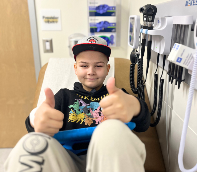 A teen boy giving two thumbs up while sitting at the doctors office for his leukemia.