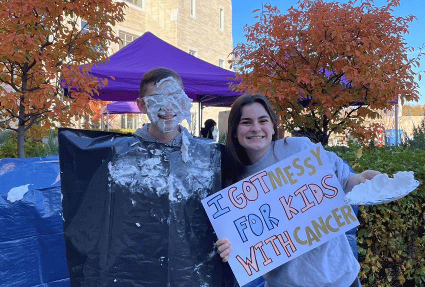 One student with whipped cream in his face and the other holds a sign that says 