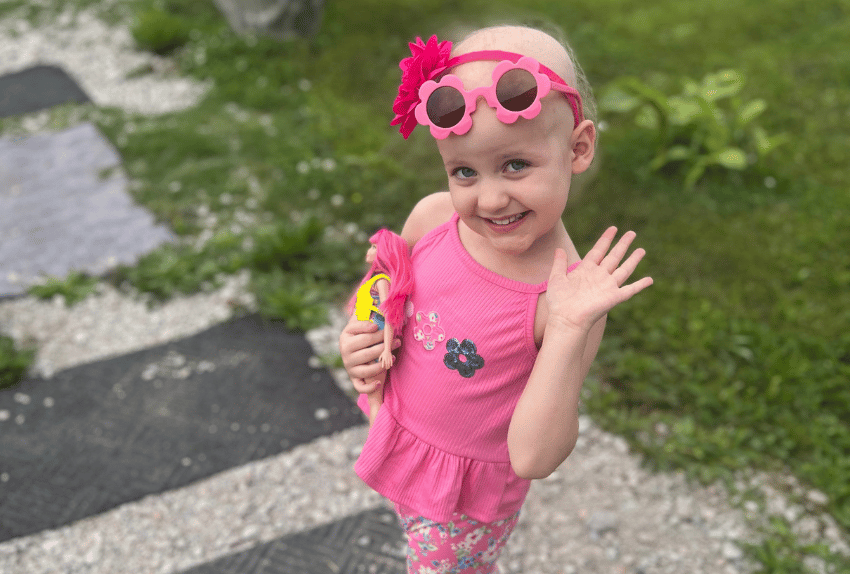 A little girl waving at the camera while holding her Barbie wearing all pink with a pink headband and pink sunglasses.
