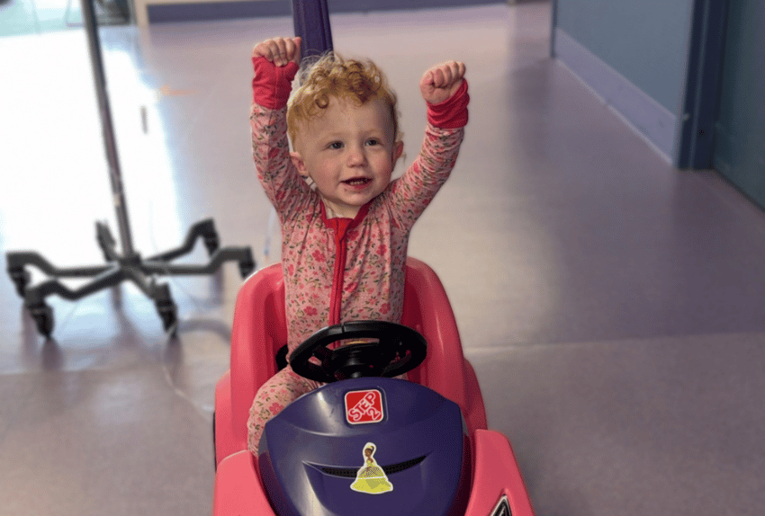 A little girl with her hands up in celebration going down the hall of a hospital on a pink car with a fluids rolling cart behind her.