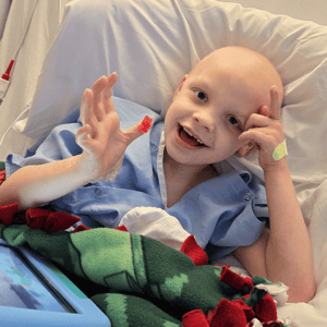 A little boy holding a gummy bear on his finger while smiling at the camera in his hospital bed, facing childhood cancer. He and his family received financial support from Pinky Swear Foundation.